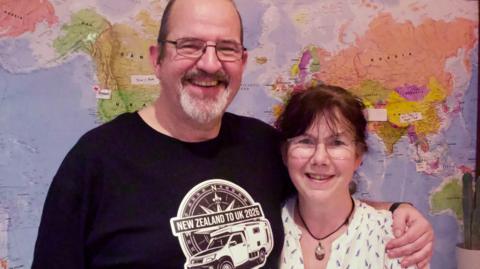 A man and woman stood in front of a wall map of the world. The man has a grey goaty beard and glasses and a black t-shirt with an image of a camper van and the words New Zealand to UK 2026. The woman is wearing a white, patterned shirt with a brown necklace. She bhas brown hair and glasses