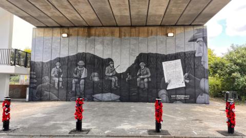 A greyscale mural painted on the wall of an underpass showing five soldiers sitting in a trench, with one writing a letter to his parents. In front of the mural are four bollards decorated with poppies.