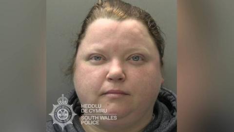 Jemma Lewis has blue eyes and brown hair. She is wearing a top with a grey collar. The top of the Gwent Police logo can be seen in this police mugshot.