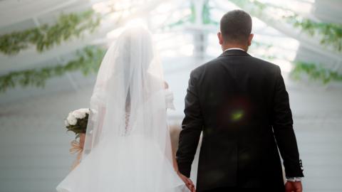 Stock photo shows the backs of a bride and groom on their wedding day, wearing a white wedding dress and black suit, the bride carries a bouquet of flowers and a marquee with flowers hanging can be seen in the background.