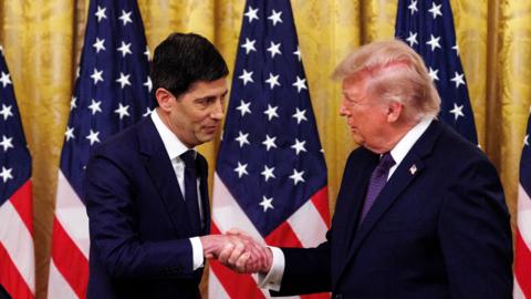 Donald Trump shakes hands with Kevin Warsh, both men wear navy suits and are flanked by US flags.
