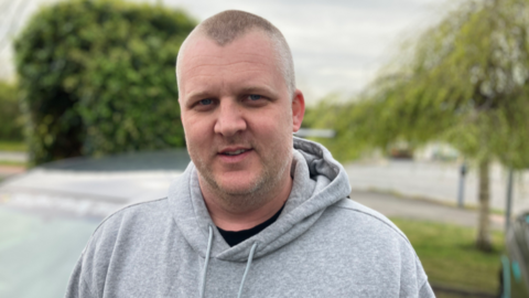A man with short hair and stubble. He is wearing a grey hoodie over a black tshirt and is smiling at the camera
