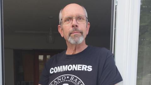 A man in his 60s stands by some patio doors. He is wearing a black T-shirt with white writing on and glasses.
