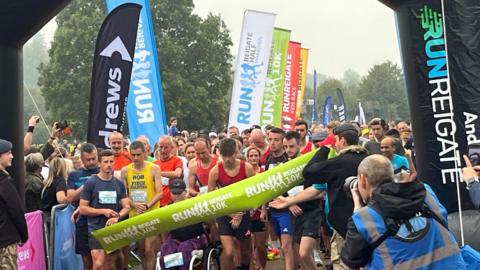 Scores of runners are lined up at the starting line, with a lime green coloured large banner running horizontally in front of them. Tall banners bearing the name of the race stand either side of the group of runners and people in the crowd hold cameras in the air and a photographer takes a photograph of the runners at the front