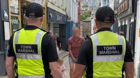 Two street marshals with their backs to the camera in a St Ives street. They both are wearing yellow hi-vis. 
