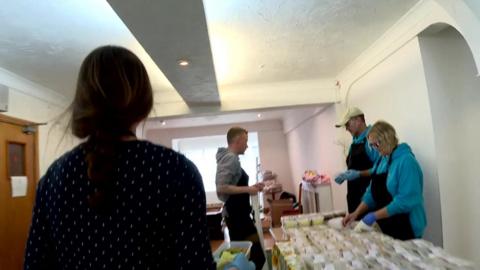 Woman walks towards a table full of meals in small plastic boxes, where three other people are working, preparing to hand out the meals