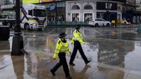Two uniformed police officers walk across a rain-soaked pavement in London, with shops, a white car, and a large lorry visible in the background.