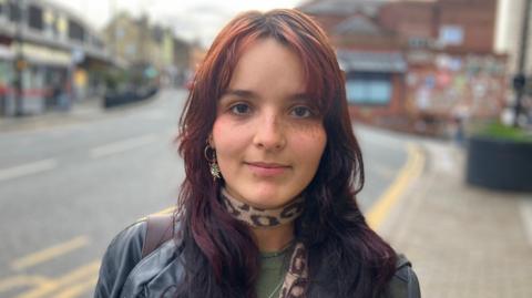 A woman with brown hair looks at the camera. She wears gold jewellery and a leopard print scarf around her neck. A bag hangs from her left shoulder and she wears a black leather jacket.