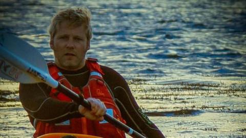 A man wearing a red life jacket sits in a kayak canoe holding a paddle, a large body of water behind him with waves and grass