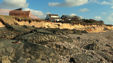 A row of large rock bags on Thorpeness beach in front of a heavily eroded coastline. In the background, there are two homes close to the edge of a low, sandy cliff. 