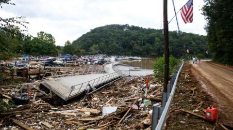 A damaged walkway - partially twisted onto its side - lies on top of debris left by Hurricane Helene in North Carolina. The walkway lies over a lake, with a road also pictured to the right of the lake.