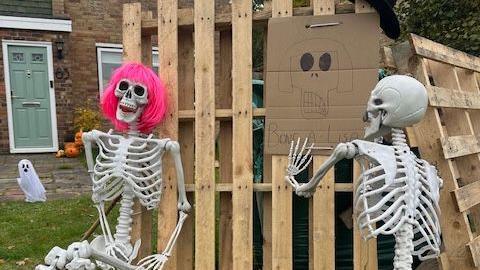 Two skeletons stood next to a piece of cardboard that says 'Bone A-Lisa'