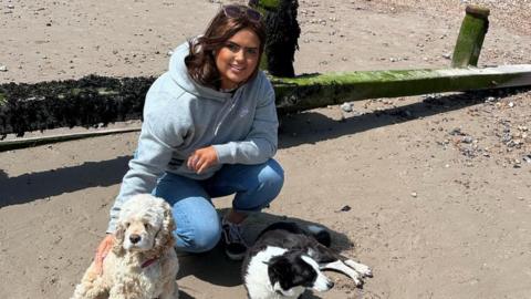 A woman crouching on the floor on a sandy beach, with two dogs sat near her. She is looking at the camera and smiling.