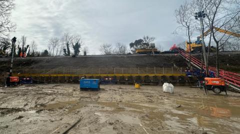 A view from the ground of where the landslip was and the embankment has been rebuilt, with machinery and people in hi-viz suits on train tracks.