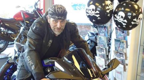 Paul Gowlett astride a black and blue motorbike in a motorbike showroom. He is wearing black bike leathers and a black bandana with white decoration. He has a light coloured beard with grey hairs. There is a magazine stand next to him with black balloons with white skulls and crossbones on them.