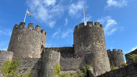 Llun o ddau dwr Castell Conwy ar ddiwrnod braf. Mae baneri ar dop y ddau dwr. 