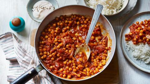 A silver pan filled with chickpeas and onion in a red sauce, on a whitewashed wooden table with a silver serving spoon.