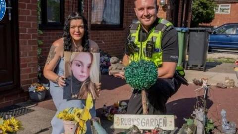 A woman and a police officer pose in front of a memorial garden. The officer is holding a small stone ornament while the woman is holding a large photo up of a young blonde teenager.
