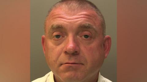 Police mugshot of a man with short hair. He is wearing a yellow shirt and is looking directly at the camera.