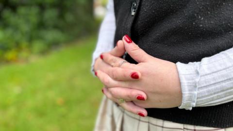 Chloe's hands pictured clasped together. She is wearing red nail polish, a long-sleeved white shirt and a black button-up vest.