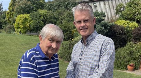 Brian Foxwell and Jamie Cox. Both men are pictured outside. They both have short light grey/white hair. Brian is wearing a blue striped shirt and Jamie is wearing a checked shirt. They are both smiling.