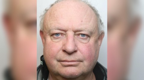 A mugshot of Brett Conway. He has light blue eyes, a little stubble grey stubble and short white hair on the sides. He is wearing a collared grey top.