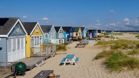 A row of brightly coloured beach huts stand on a sandy beach next to some small dunes. There are picnic benches outside the huts. The sky is blue and it is a sunny day.