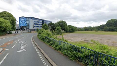 King's Road in Plymouth. There is a large blue and grey building on the left and a grassy field on the right of the road.