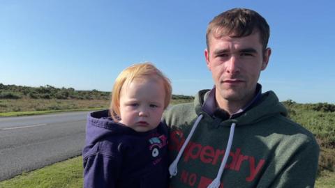 Dan Rangecroft, standing alongside a road in the New Forest, holding his 16-month-old daughter