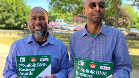 Two men stand smiling at the camera. they are outside and each holding a poster which reads 7th Suffolk BME Business Awards.