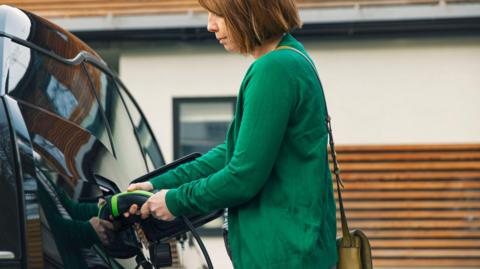 Woman wearing a green jumper plugging a charging cable into a black electric vehicle