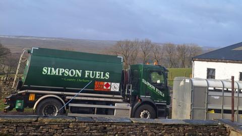A Simpson Fuels tank is parked up outside the end of a white house which is partly obscured by a horsebox. 