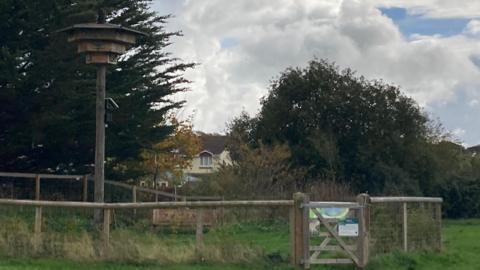 Picture shows a wooden tower with bird box at the top, and a wooden fence around a pond. There is a wooden bench for people to sit on