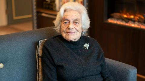 Vera Schaufeld dressed in black sits on a sofa. She has a brooch on her jumper