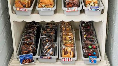 A selection of cakes, in clear plastic packaging, sit in trays across two white shelves.