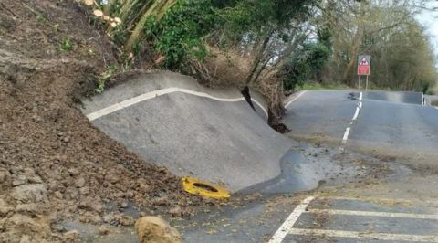 One side of a road has been pushed up by a landslip under it and it resembles a skate ramp. In the distance, a large dip can be seen in another section of the route alongside a large crack in the road's surface.