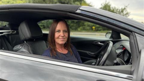 Caroline O'Brien sitting in her car looking directly at the camera. She is wearing a blue top. She is visible from the shoulders up and is sitting in the driver's seat with the steering wheel in front of her. There is a road and foliage in the background.