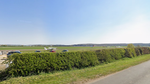 A relatively empty and large field with a few cars and a small white plane in it. In the foreground is a hedge and a strip of grass next to a road