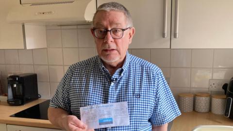 David Lane has grey hair. He is wearing black-framed spectacles on his face and a blue-checked shirt. He is holding up his Yorkshire Water bill while standing in his kitchen in front of the hob.