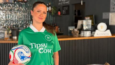 Freya, a woman with brown hair, wearing a green jersey holding a white and blue football, smiling for camera.