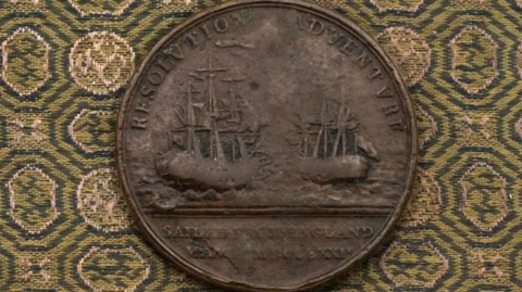 A medal with images of the ships used by Captain James Cook on his second voyage. The medal is sitting on a green and yellow patterned cloth.