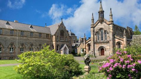 Several historic buildings with a green garden in front and pink flowers 