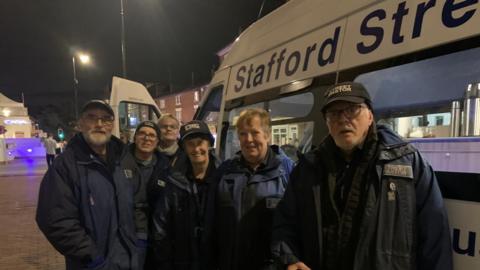 Six people are standing beside a white van that says "Stafford Street Pastors". They are all in dark coats and some have black caps that read "street pastor"