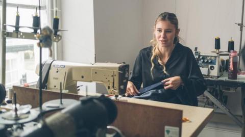 Molly Haney is sitting at a sewing machine using a needle and thread to sew together a piece of fabric. She is surrounded by sewing machines and other tailoring apparatus.