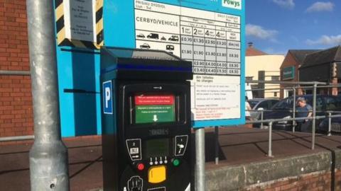 A black parking meter, with a sign listing charges behind it and a lampost to the left.