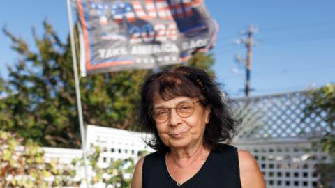 June in her garden, looking at the camera smiling, in front of a white fence with plants, a blue sky and a flag printed with the words Trump 2024. She is wearing a black cropped top and round wireframe glasses.