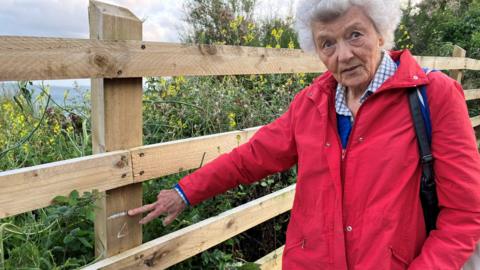 An older lady with grey hair and a red coat points to a white mark she has drawn on a fence post to mark the original height of the fence at 90 cms. 