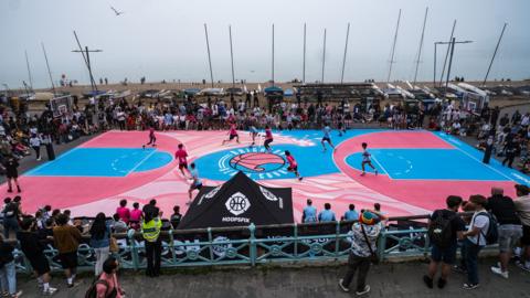 People playing on a blue and pink basketball court with a pebble beach and the ocean in the background.