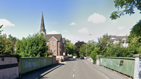 A green bridge with white graffiti on the side of Rock Lane West with a block of flats in the distance