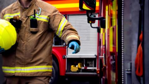 A firefighter, his face and head just out of shot, is carrying a yellow fire helmet and with one hand reaches out towards the door of a fire engine.